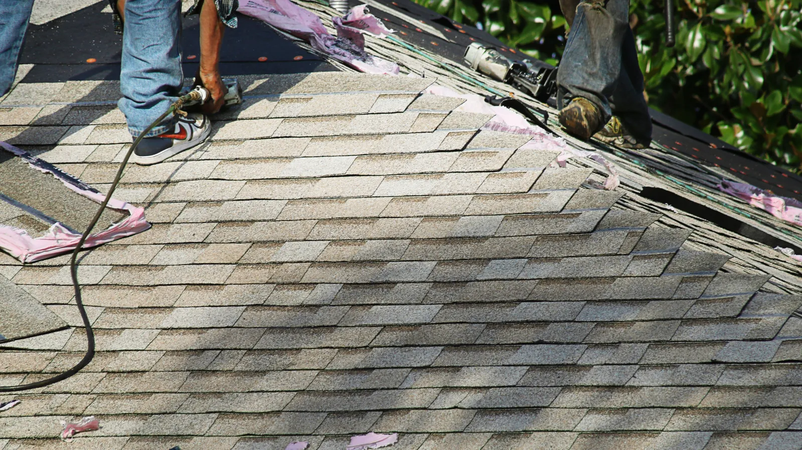 Near the ridge a roofer uses a nail gun on light brown shingles